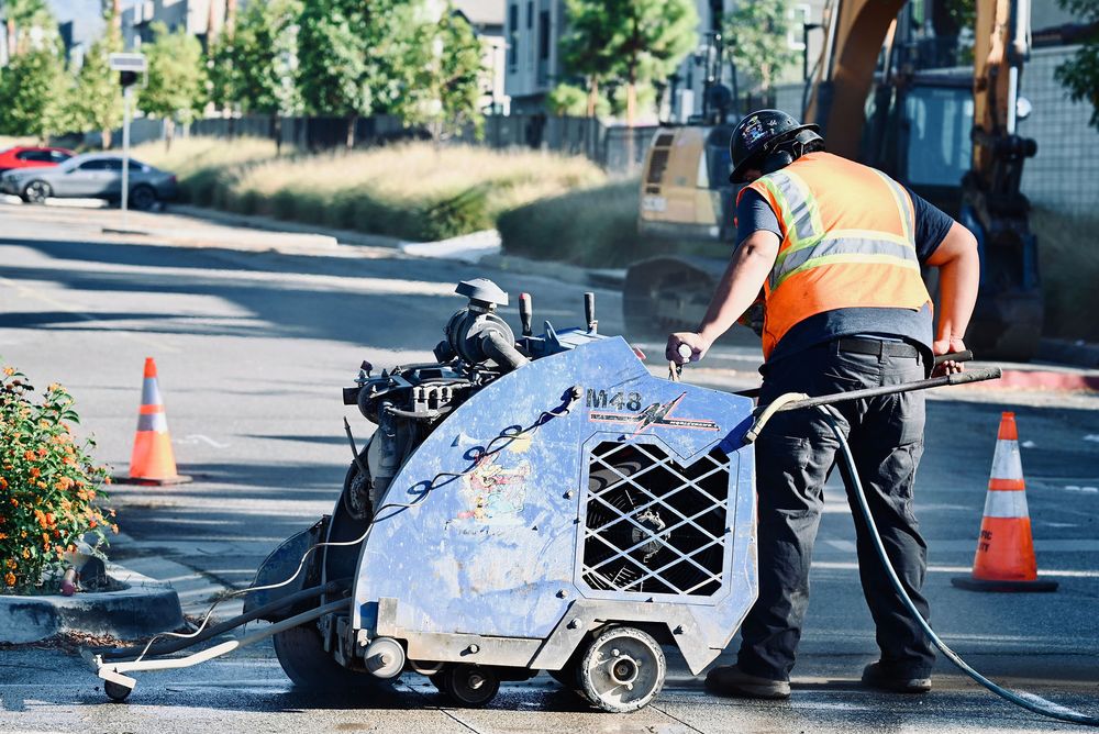 Operator flat-sawing a curb with an excavator on the job behind him
